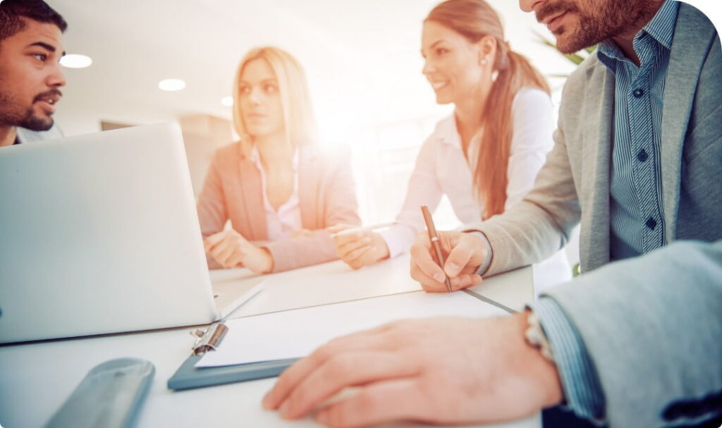 Gruppo di professionisti riuniti attorno a un tavolo durante una riunione di lavoro in un ufficio luminoso, con laptop e documenti