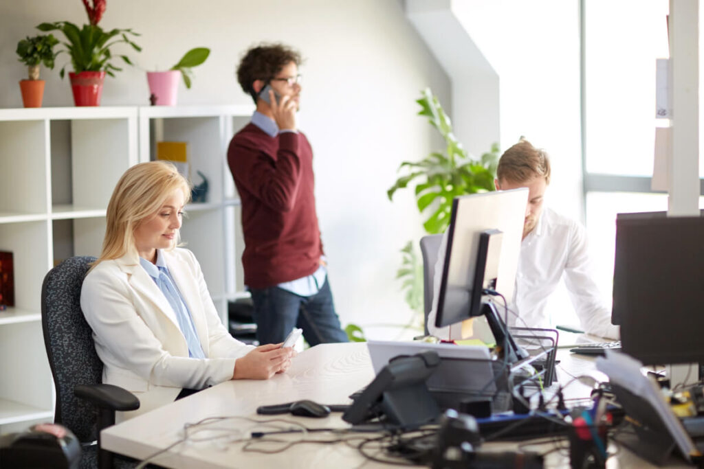 Team di professionisti al lavoro in un ufficio luminoso e accogliente: una donna in primo piano consulta documenti alla scrivania, un collega parla al telefono in piedi e un altro lavora al computer