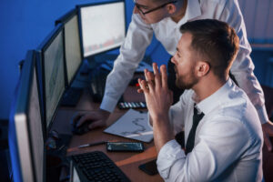 Due professionisti in camicia bianca analizzano dati su monitor multipli in un ufficio con illuminazione blu, con documenti e grafici sulla scrivania