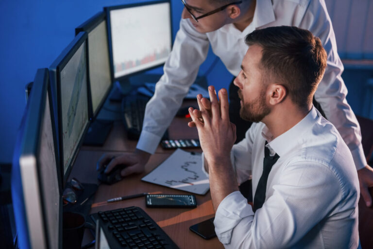 Due professionisti in camicia bianca analizzano dati su monitor multipli in un ufficio con illuminazione blu, con documenti e grafici sulla scrivania