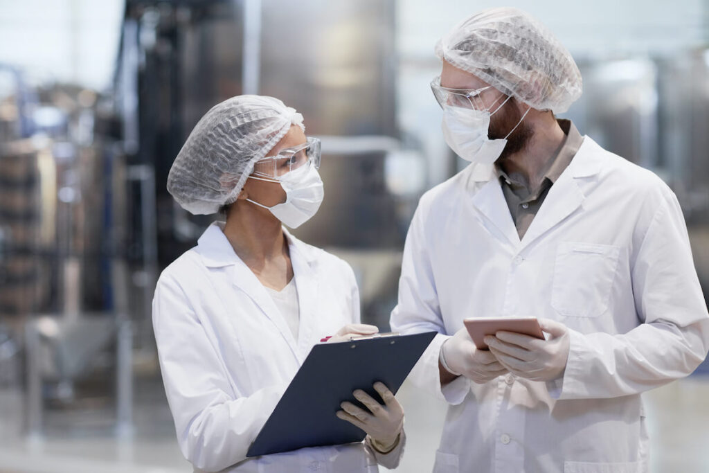Due operatori in camice bianco, cuffia e mascherina consultano una clipboard e un tablet all'interno di un impianto di produzione alimentare