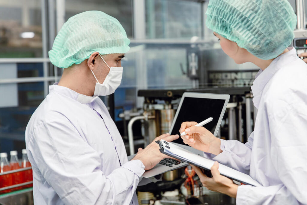 Due operatori con cuffia verde e camice bianco verificano dati su clipboard e laptop all'interno di un impianto di produzione alimentare