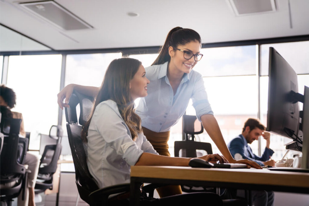 Due colleghe sorridenti collaborano davanti a un monitor in un ufficio open space luminoso, con altri colleghi sullo sfondo