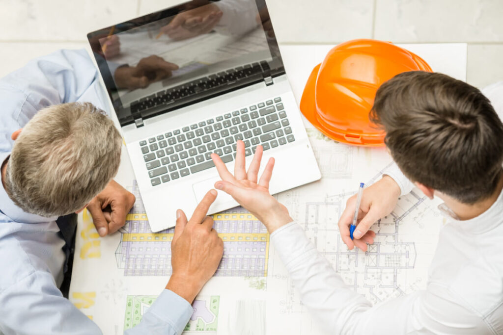 Due professionisti lavorano su planimetrie tecniche con un laptop aperto su un tavolo, con un elmetto arancione posato accanto