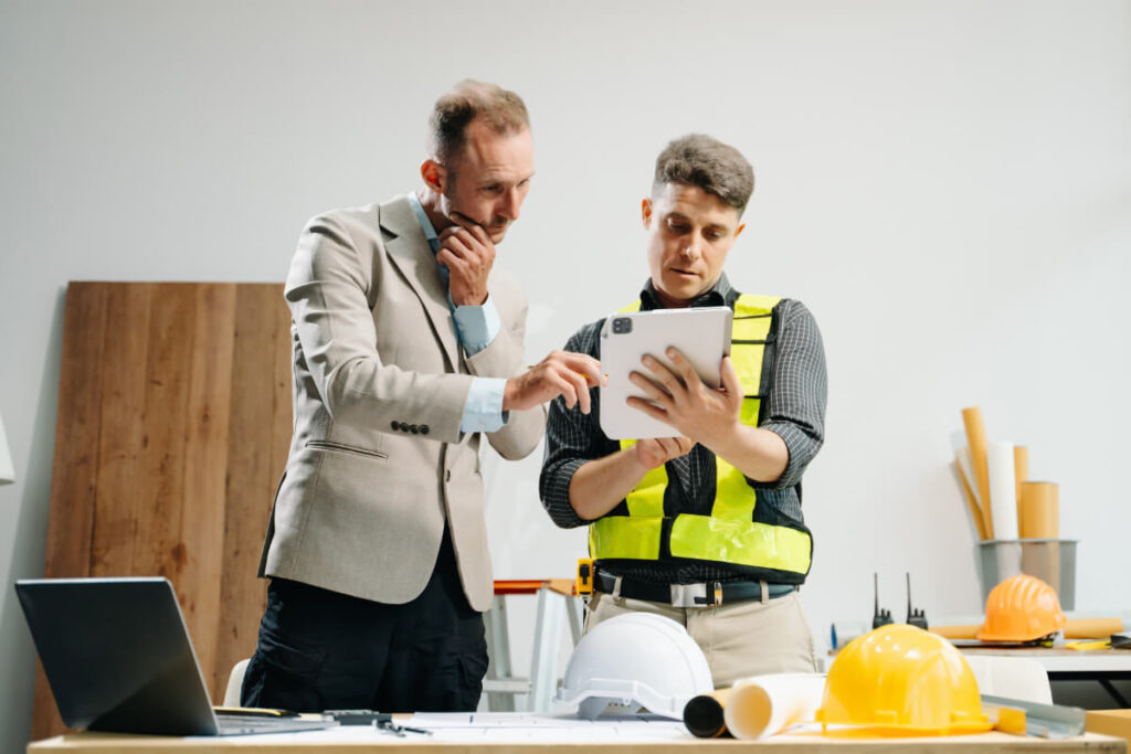 Un manager in giacca e un tecnico con gilet catarifrangente esaminano insieme documenti su un tablet, con elmetti e materiali da cantiere sullo sfondo