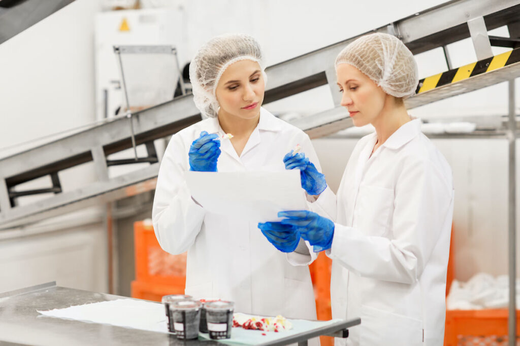 Due operatrici con cuffia, camice bianco e guanti blu eseguono controlli su campioni alimentari consultando un documento in un ambiente di produzione