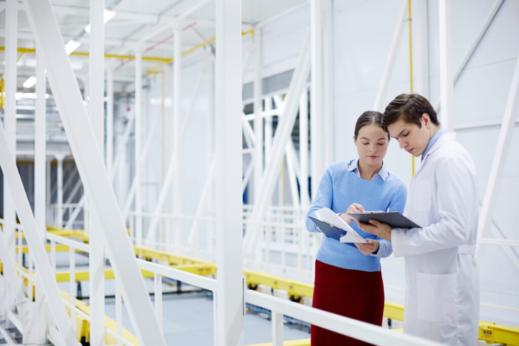 Due tecnici consultano appunti su una clipboard all'interno di un impianto industriale con strutture metalliche bianche e gialle