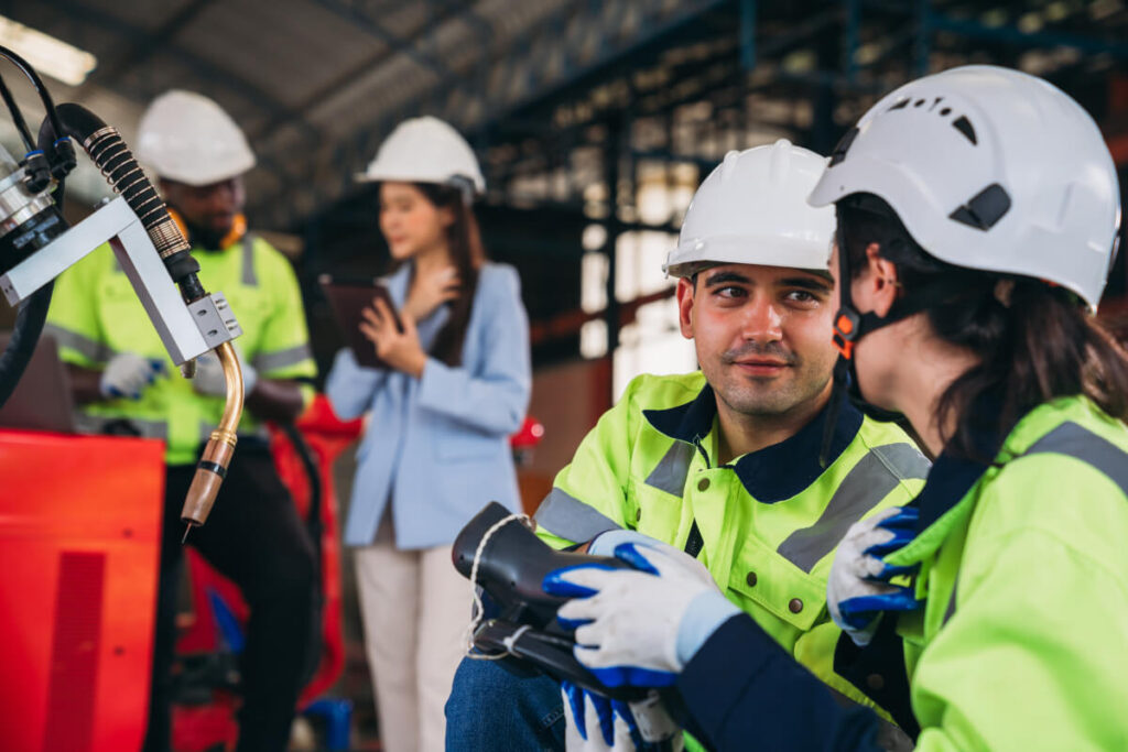 Due operatori in primo piano con elmetto bianco e gilet catarifrangente discutono accanto a macchinari industriali, con altri colleghi sullo sfondo