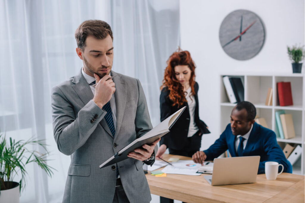 Un manager in primo piano consulta pensieroso un raccoglitore di documenti, mentre due colleghi lavorano sullo sfondo in un ufficio